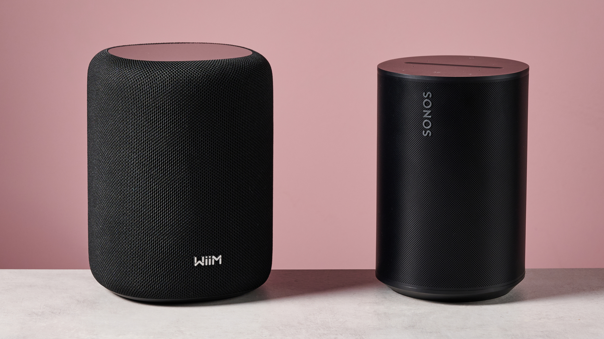 The WiiM Sound Lite side by side with the Sonos Era 100 in front of a pink background.