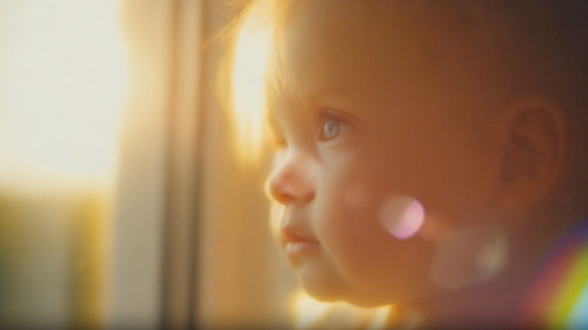 A close-up side profile of a baby's face looks towards a bright window, with soft golden light and lens flares illuminating the scene.