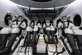 four astronauts in a crew dragon. the astronauts are seated in white spacesuits