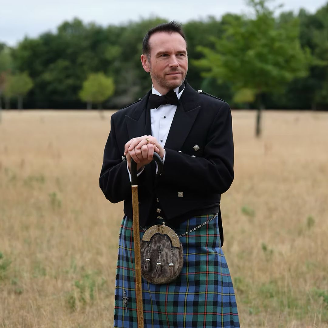 Grant Harrold wearing a bow tie and jacket with a kilt, standing in a field holding a walking stick