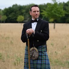 Grant Harrold wearing a bow tie and jacket with a kilt, standing in a field holding a walking stick