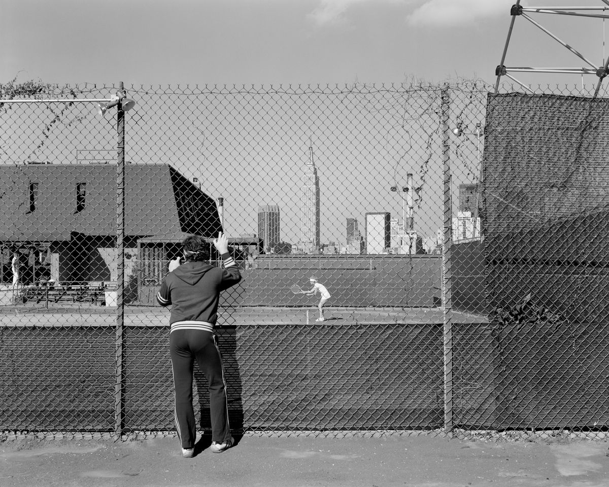 A man in a tracksuit grips a chain-link fence to watch a tennis match, with the Manhattan skyline visible beyond the court.