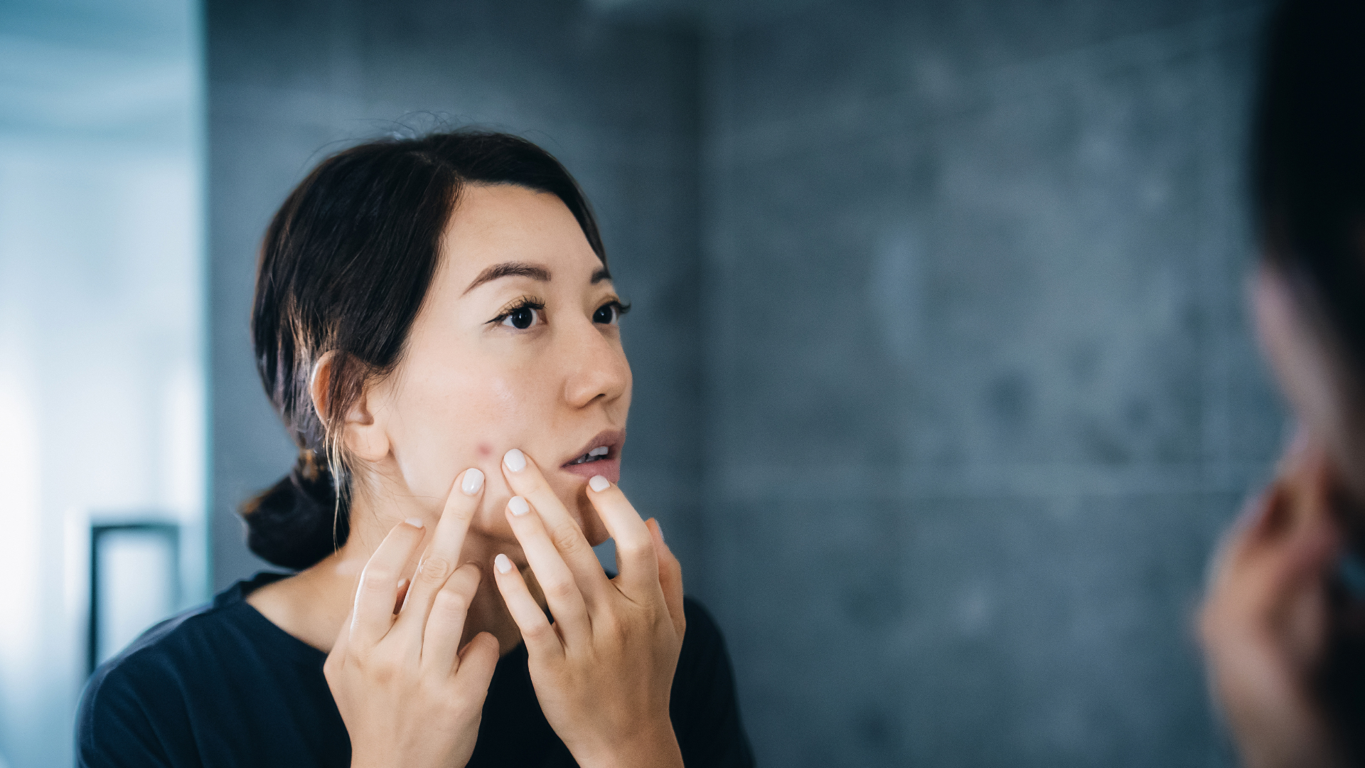 A woman looks in the mirror touching her skin around a visible pimple.
