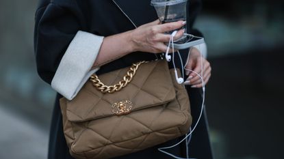 A woman carrying a brown quilted chanel bag, an iced coffee and her phone with headphones, wearing a chrome autumn nail trend