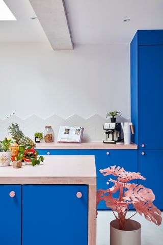 A kitchen with blue cabinetry, wooden island with blue doors, fruit bowl on island, pink palm houseplant. In background, coffee machine, recipe book on stand.
