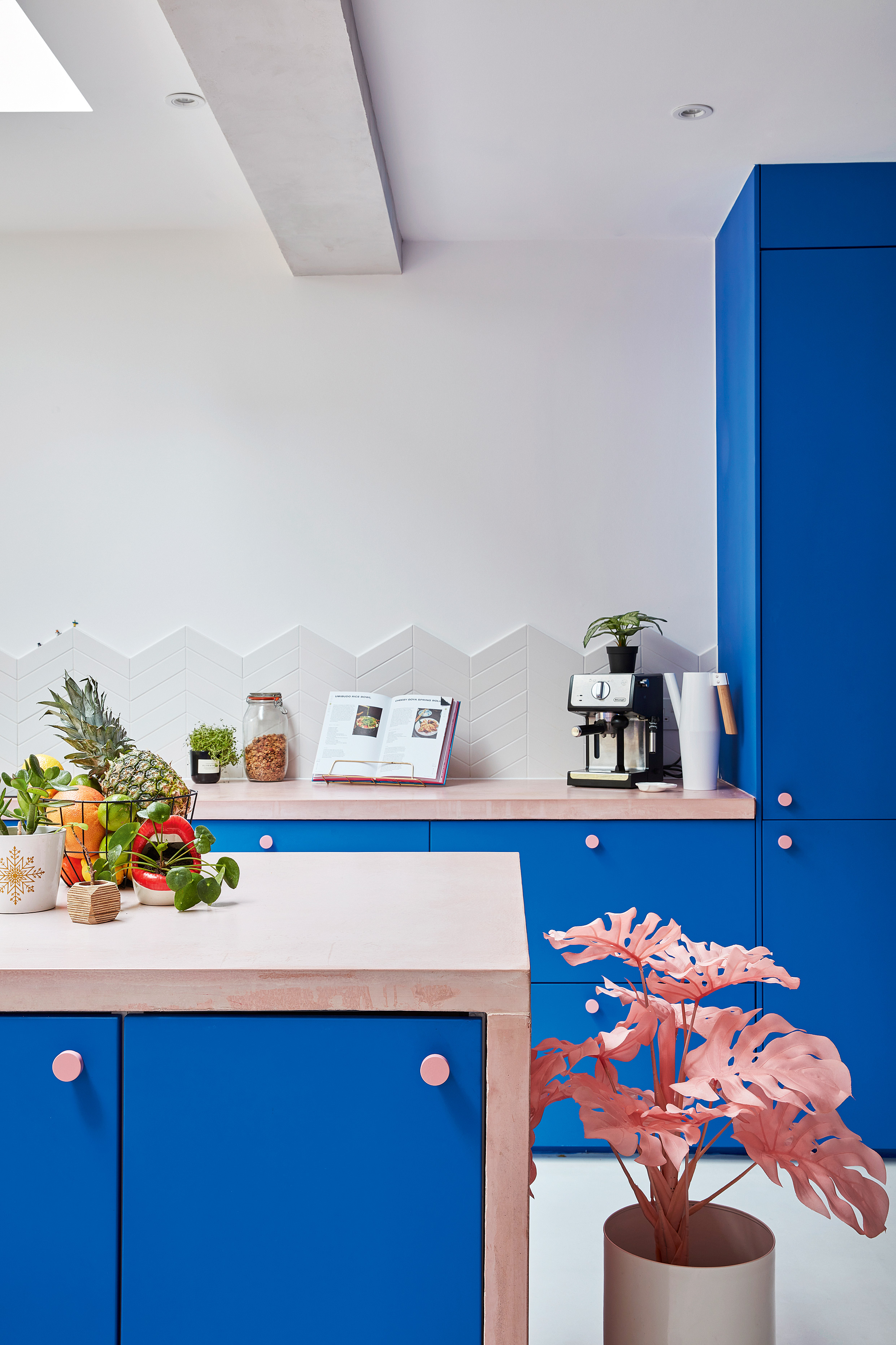 A kitchen with blue cabinetry, wooden island with blue doors, fruit bowl on island, pink palm houseplant. In background, coffee machine, recipe book on stand.