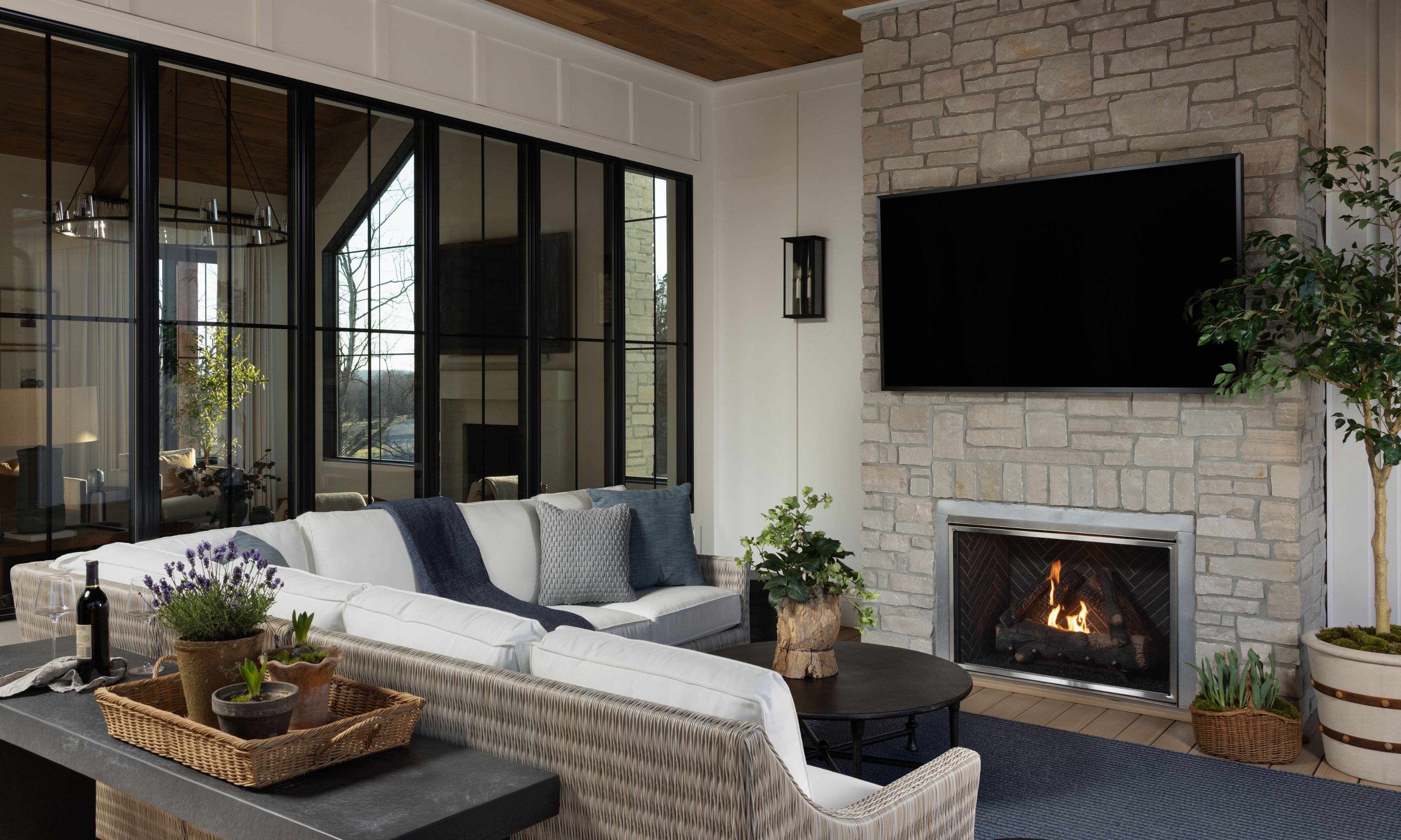 Cozy outdoor living room featuring a woven white sectional, stone stacked fireplace with mounted TV, and large glass windows overlooking nature.