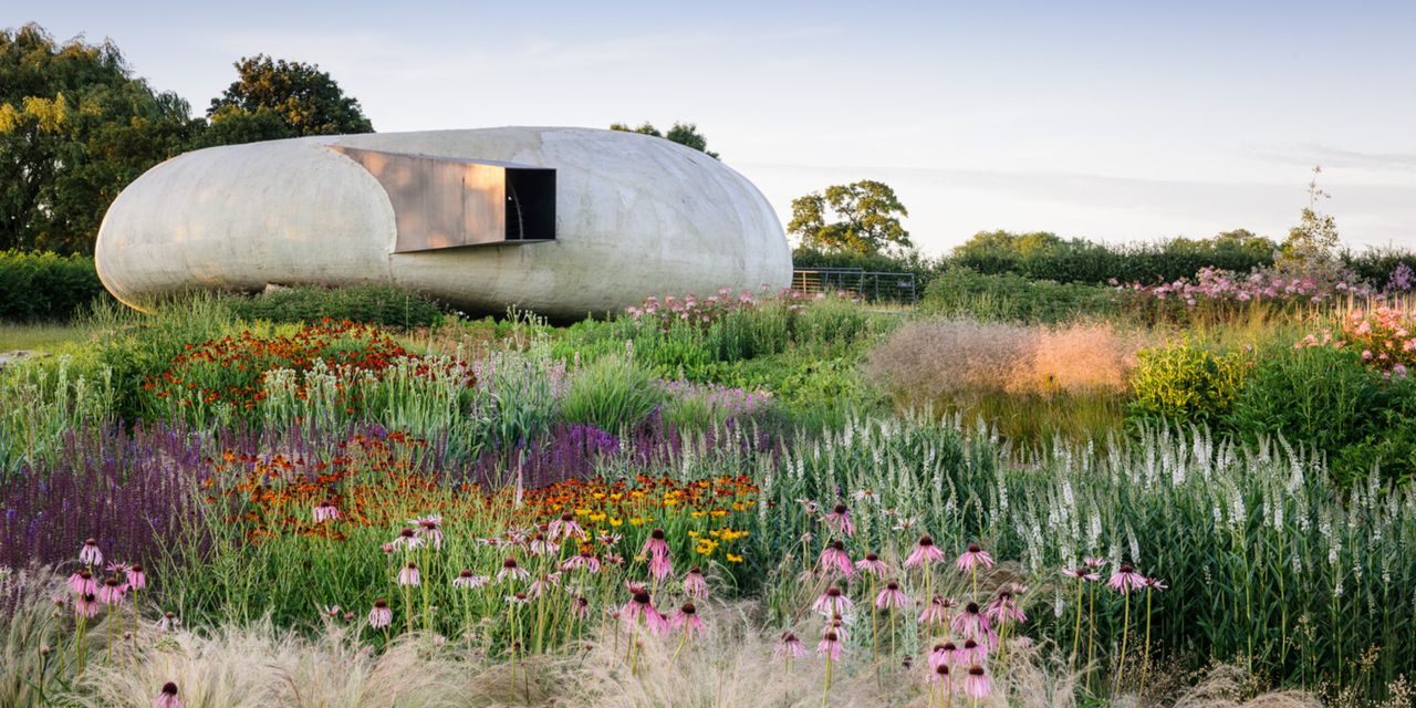 A flowers-filled field features bright patches of green, purple, red, pink, and orange, in front of a futuristic, oblong cement building at sunset.