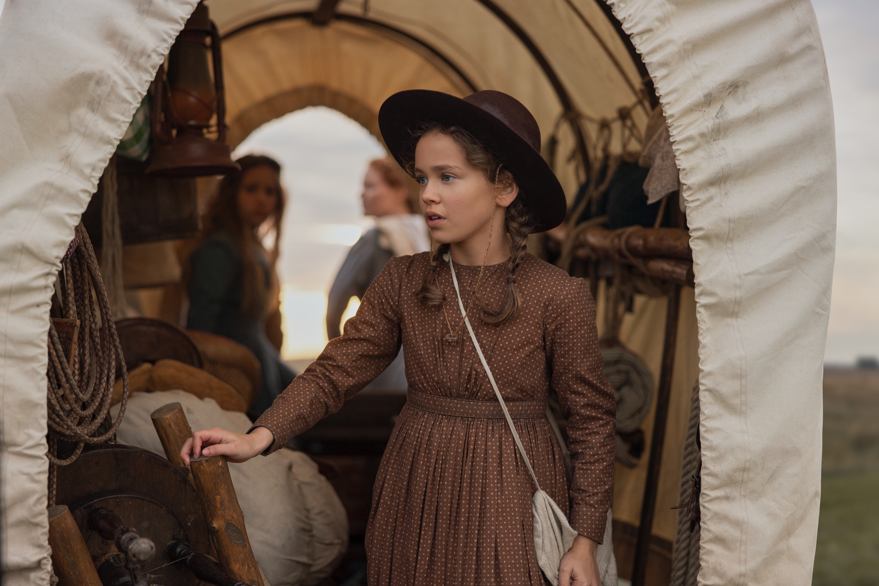 Alice Halsey as Laura Ingalls wears frontier-era clothing while standing in front of a covered wagon Her mother and sister are seen in the background, in Netflix's 'Little House on the Prairie.'