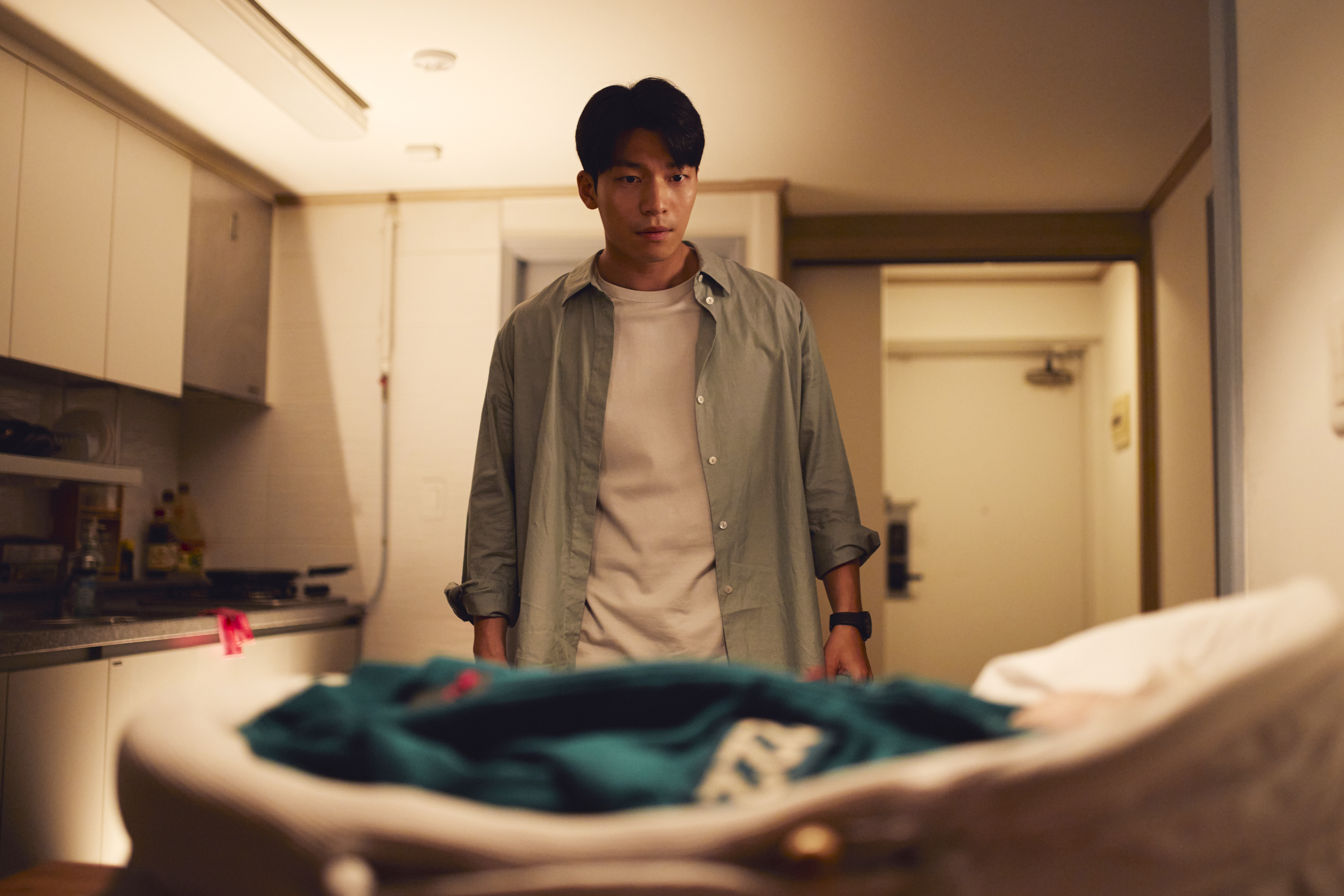 A man (Wi Ha-jun) looks at a baby, wearing a green 222 jacket, in a bassinet while standing in an apartment, in 'Squid Game' season 3.