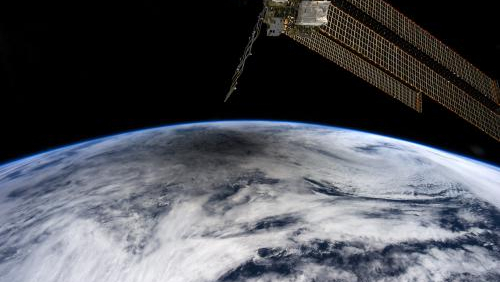 A round shadow falls on the top of a rounded Earth from space, Solar panels from the space station protrude from above.