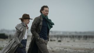 Claire Danes as Cora Seaborne in a hat and long coat and Tom Hiddleston as Will Ransome in a dark jacket and green scarf on the beach in The Essex Serpent