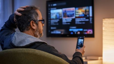 Mature man on a couch at home, holding a smartphone and watching a smart TV