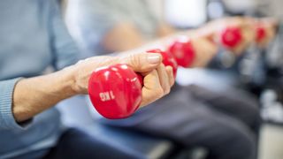 close up on an older woman's hand as she lifts a small red dumbell in a gym