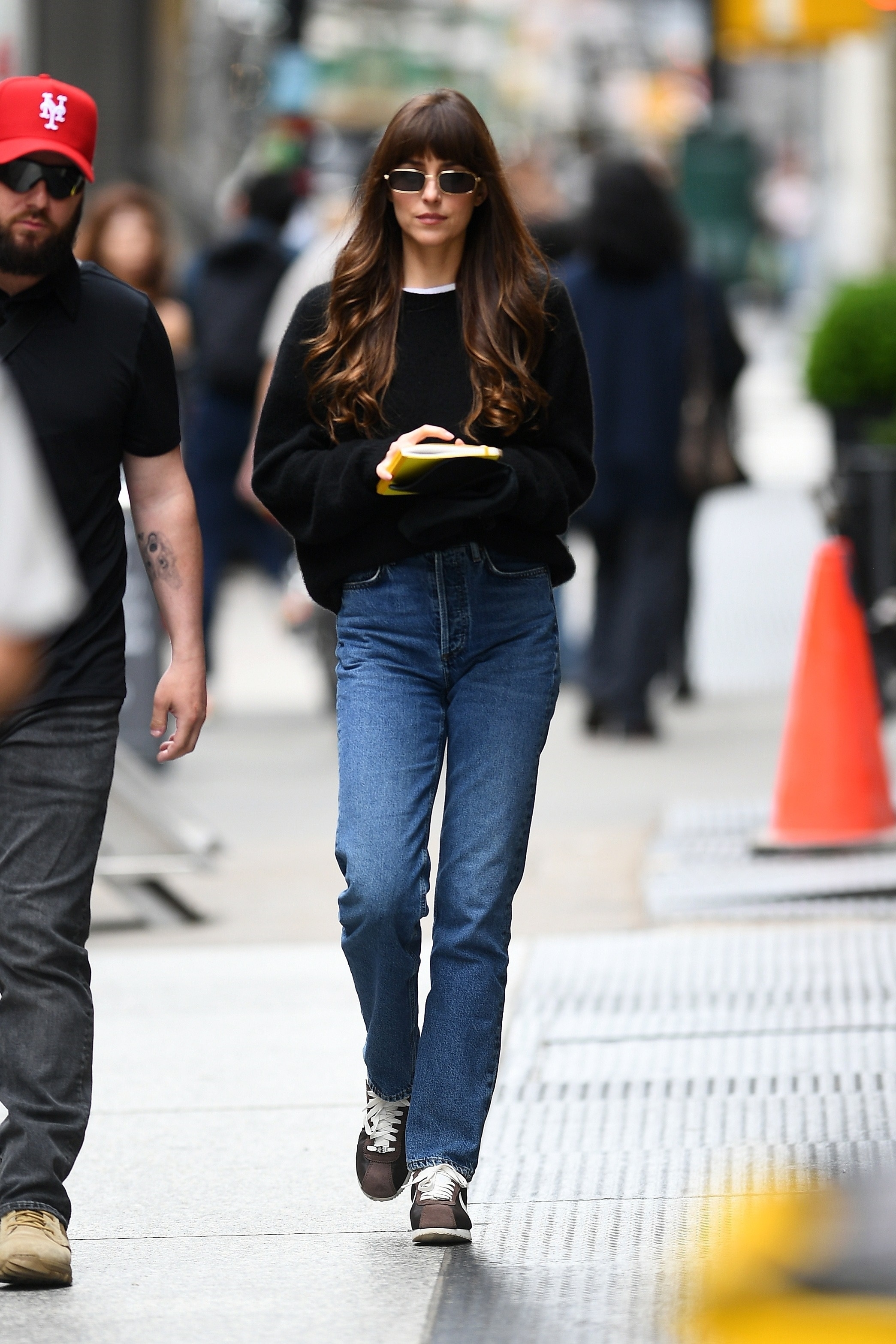 Dakota Johnson walking in New York City wearing a black sweater, dark-wash jeans, and brown Nike Cortez sneakers