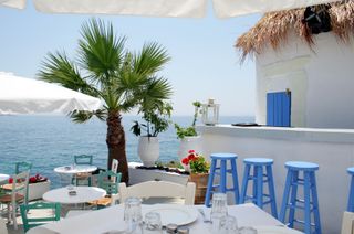 Greek taverna with white dining table in the foreground and blue stools at a bar behind. In the background, there are more dining tables and views of a palm tree and sea.