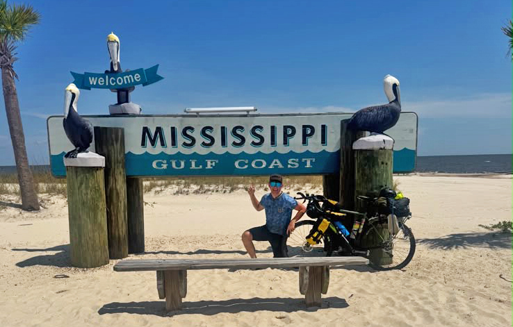 Adventure cyclist poses for photo by Mississippi sign