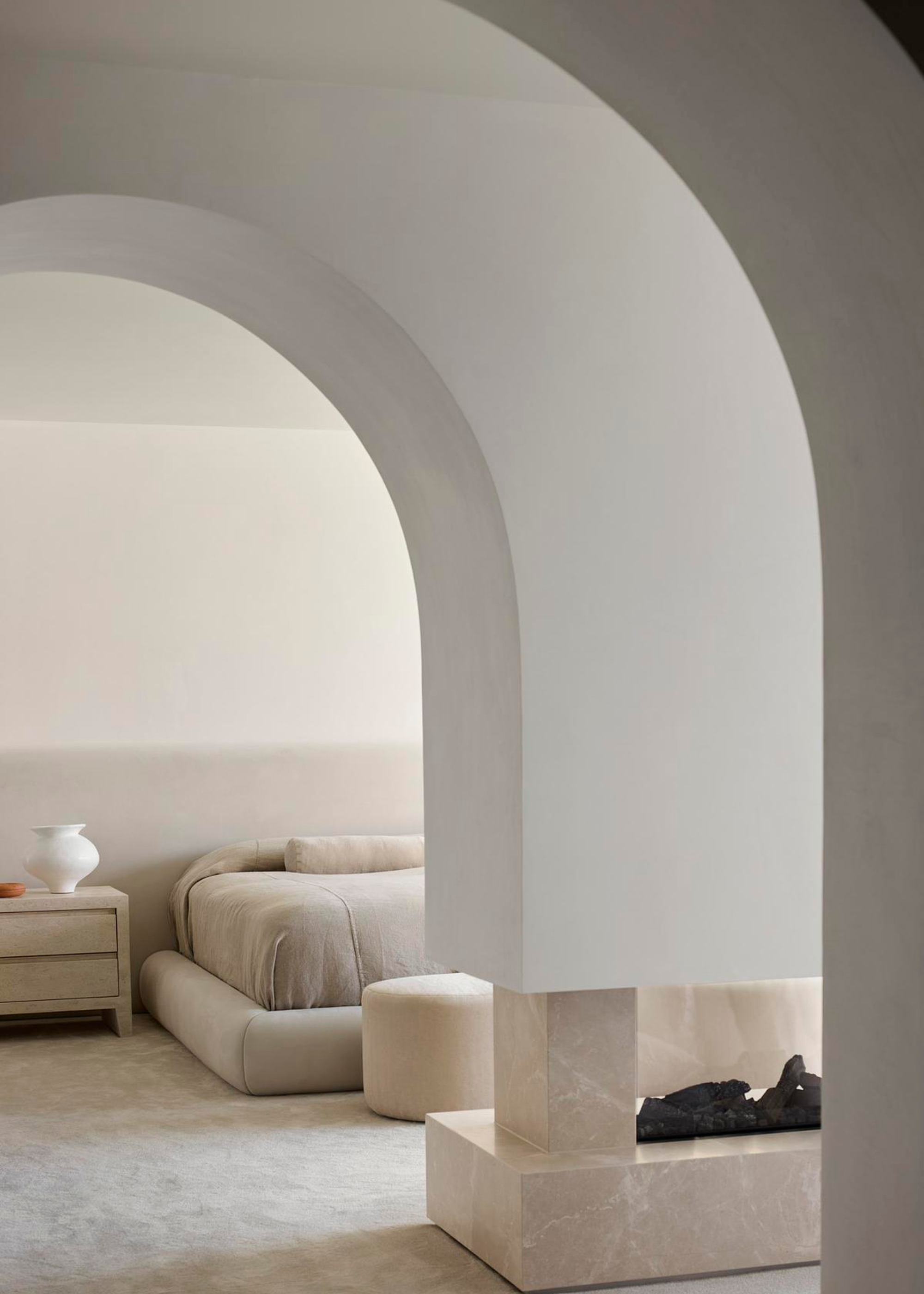 An arched doorway leading to a beautiful minimalist white bedroom with a side table holding a vase, a low-bed with a pouf and a fireplace