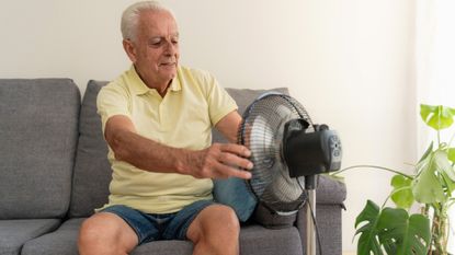Elderly man cooling off with electric fan while sitting on sofa at home during summer heatwave