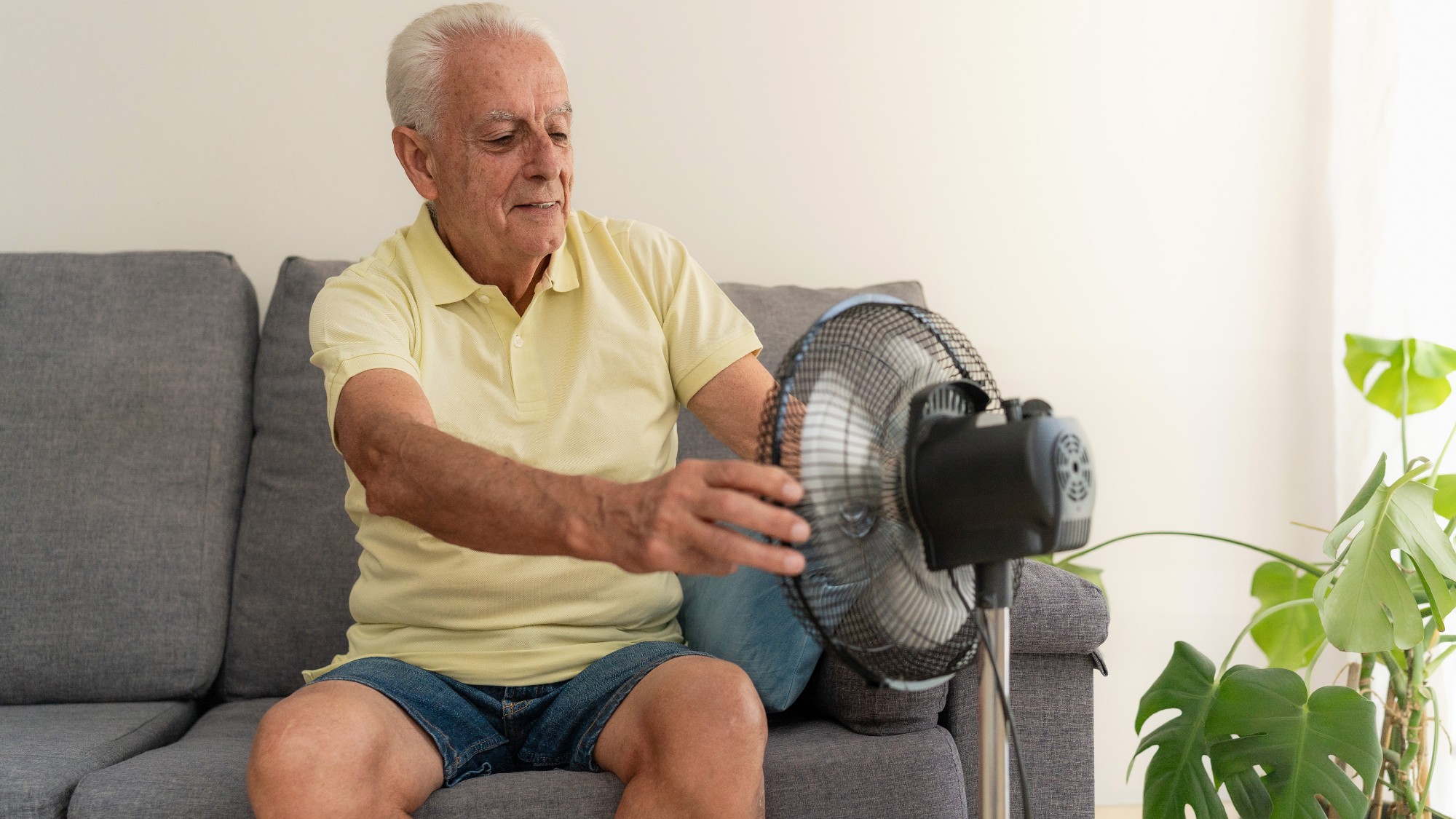 Elderly man cooling off with electric fan while sitting on sofa at home during summer heatwave