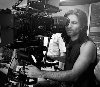 a man in a black tank top peers through the eyepiece of a large movie camera
