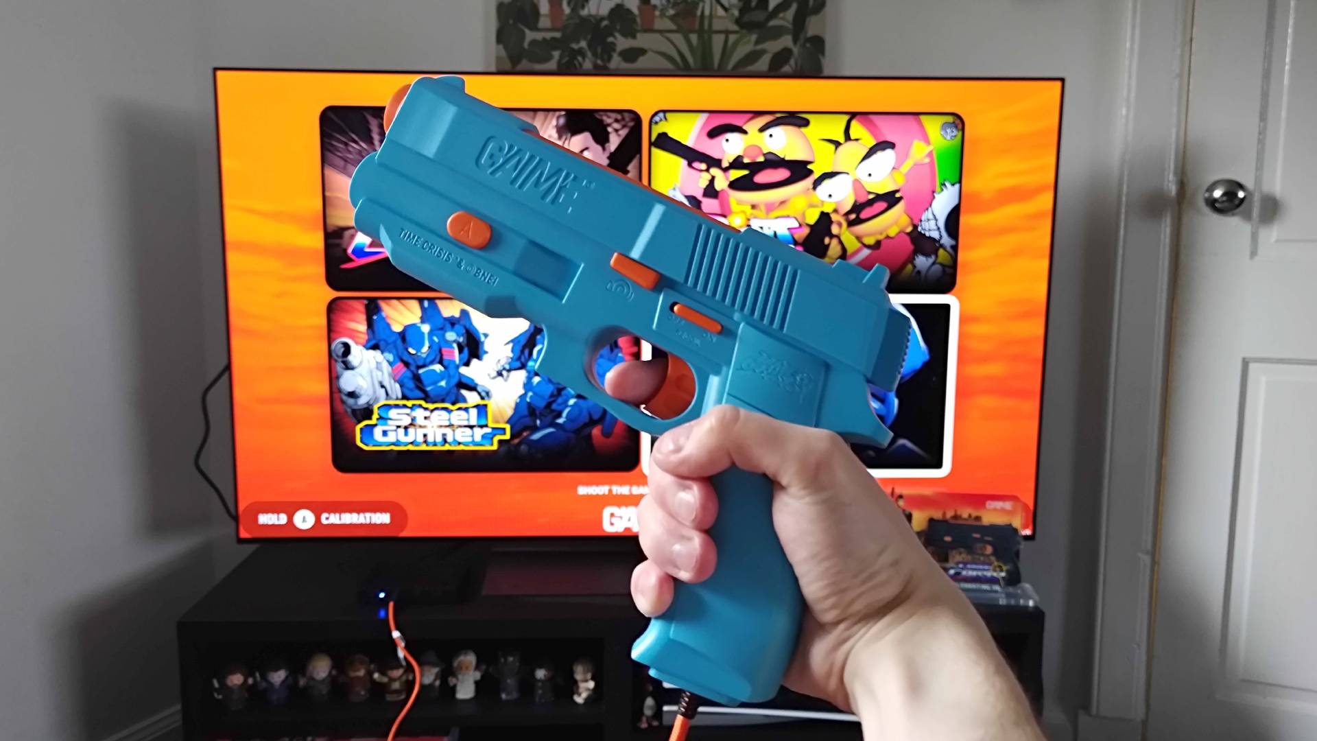 Hand holding G&#039;AIM&#039;E Time Crisis Lightgun in front of TV with game menu displayed.