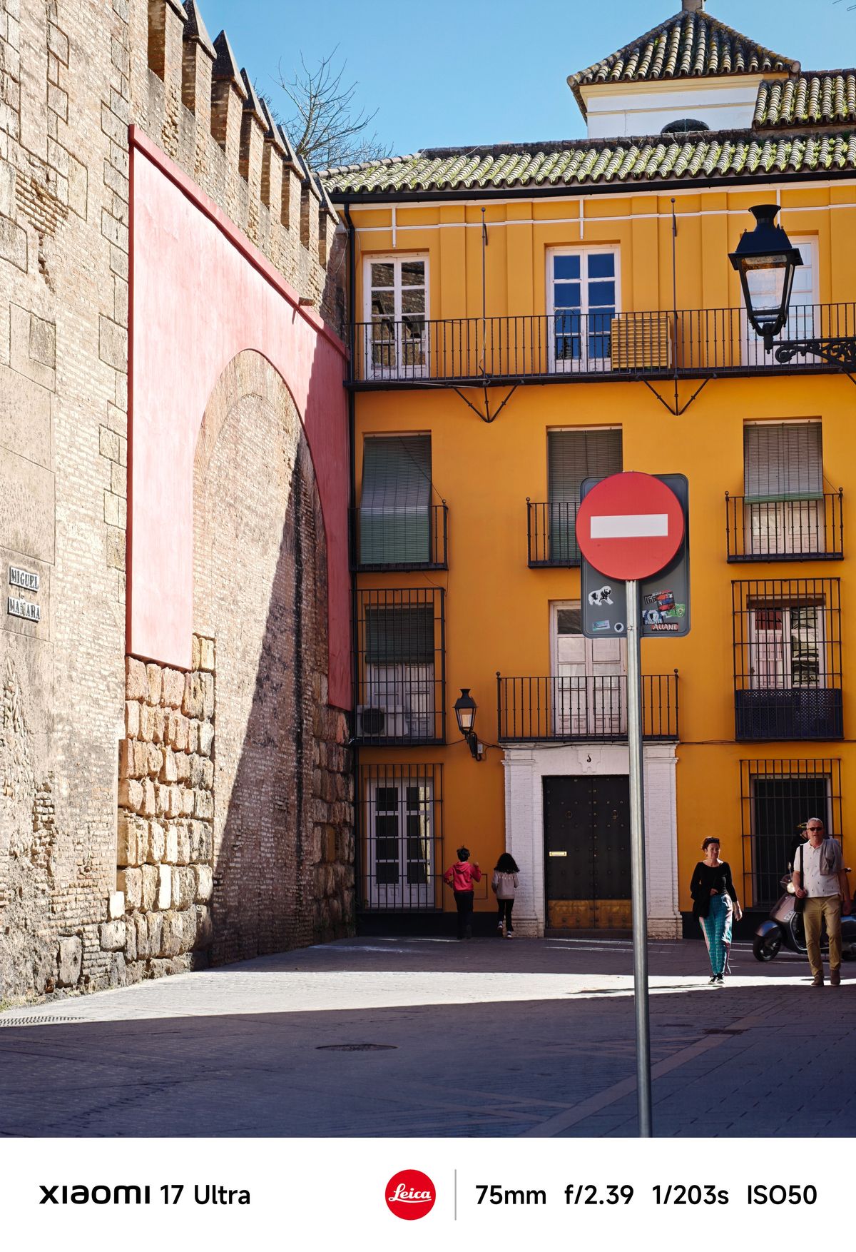 Closer view of a yellow townhouse beside a textured stone wall, red no-entry sign in the foreground and pedestrians passing by.