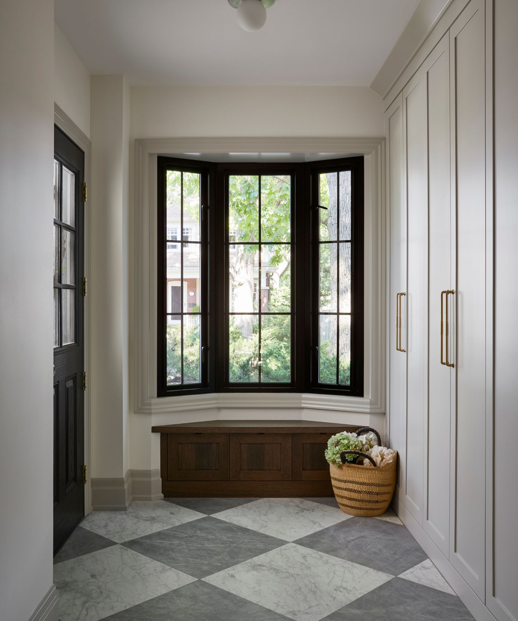 a bright entryway with a large bay window and dark wooden window seat, grey and white marble checkerboard floor, and custom mudroom joinery
