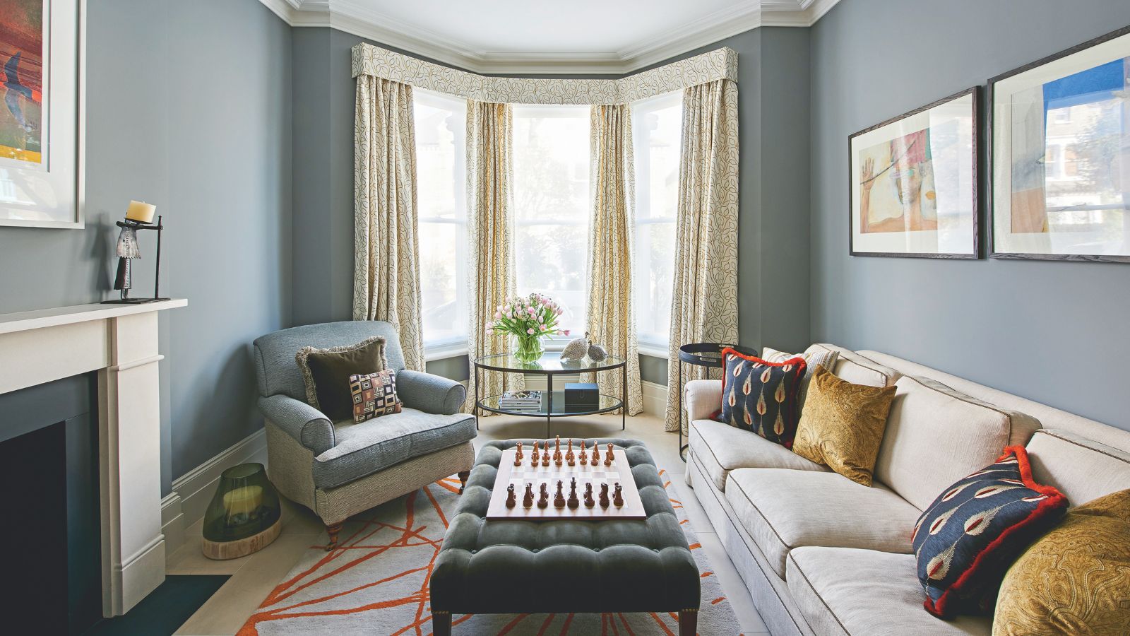 A blue living room with large bay windows with yellow curtains, a large cream couch with cushions, and a blue upholstered chair. In the centre of the room under the windows is a large circular glass table with pink tulips on it, and in the foreground is a green velvet ottoman on top of a patterned rug with a wooden chessboard on top. To the left is a white mantelpiece.