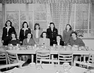 A black and white photo of ten women, with five standing behind five seated at a table.