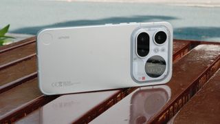 A light gray smartphone resting horizontally on a wet, reflective wooden bench. The back of the phone is visible, showcasing the camera island, the circular dot-matrix display, and a small red decorative square.