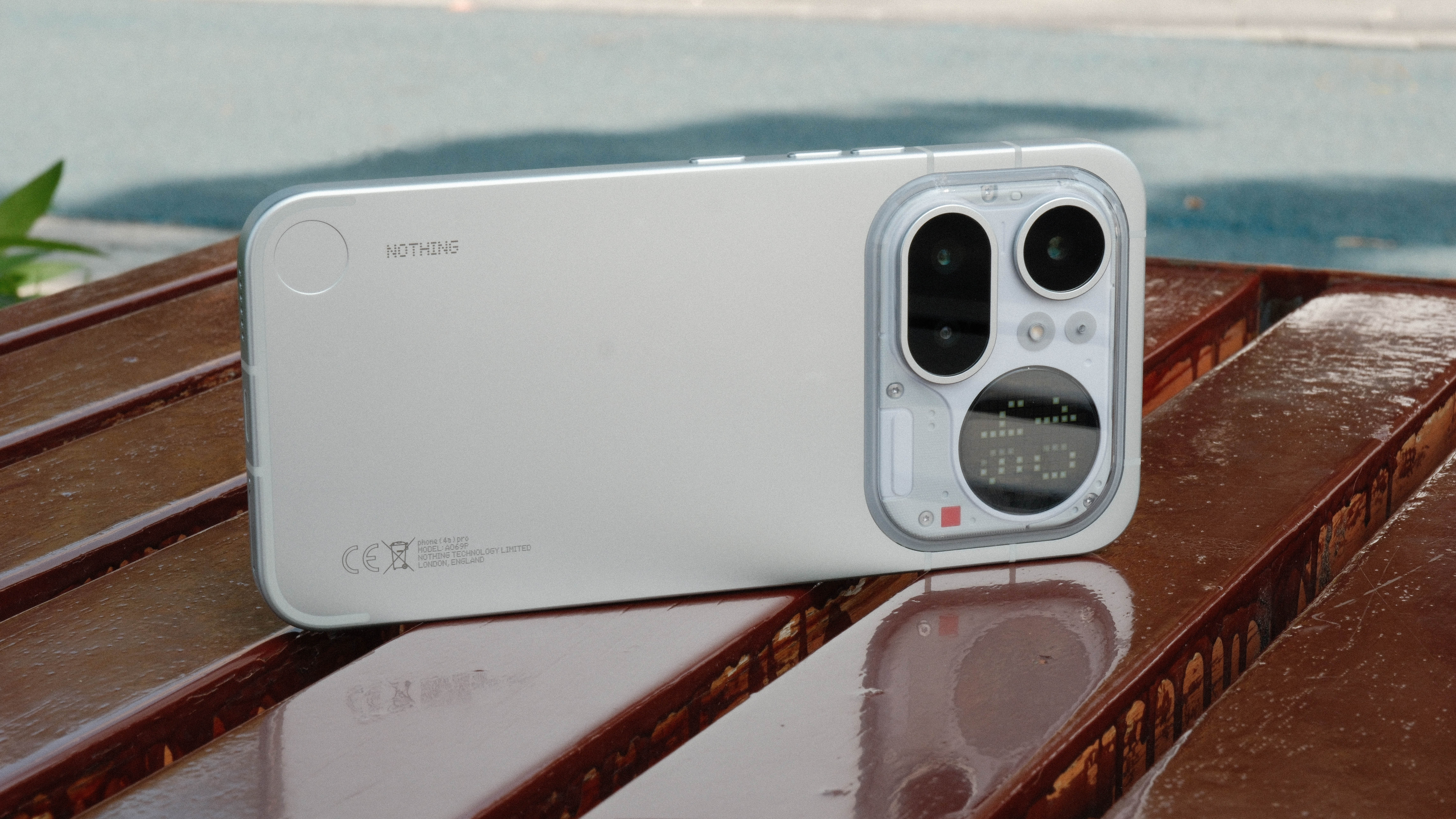 A light gray smartphone resting horizontally on a wet, reflective wooden bench. The back of the phone is visible, showcasing the camera island, the circular dot-matrix display, and a small red decorative square.