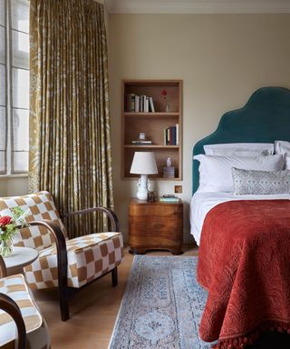 A bedroom with an inset niche filled with book above a bedside table. Beside a bed with a large blue fabric headboard and a red throw blanket