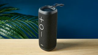 a black oblong-shaped bluetooth speaker with mesh covering and a carry strap photographed on a beige table in front of a navy wall