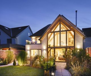 A renovated self build home lit up in the evening with wall lights on the exterior and uplit garden plants