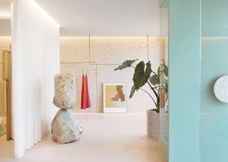 In a fashion boutique, artworks including a totemic stone sculpture and a softly colored painting appear side by side with hanging white curtains and large plants in a gold-shaded environment.