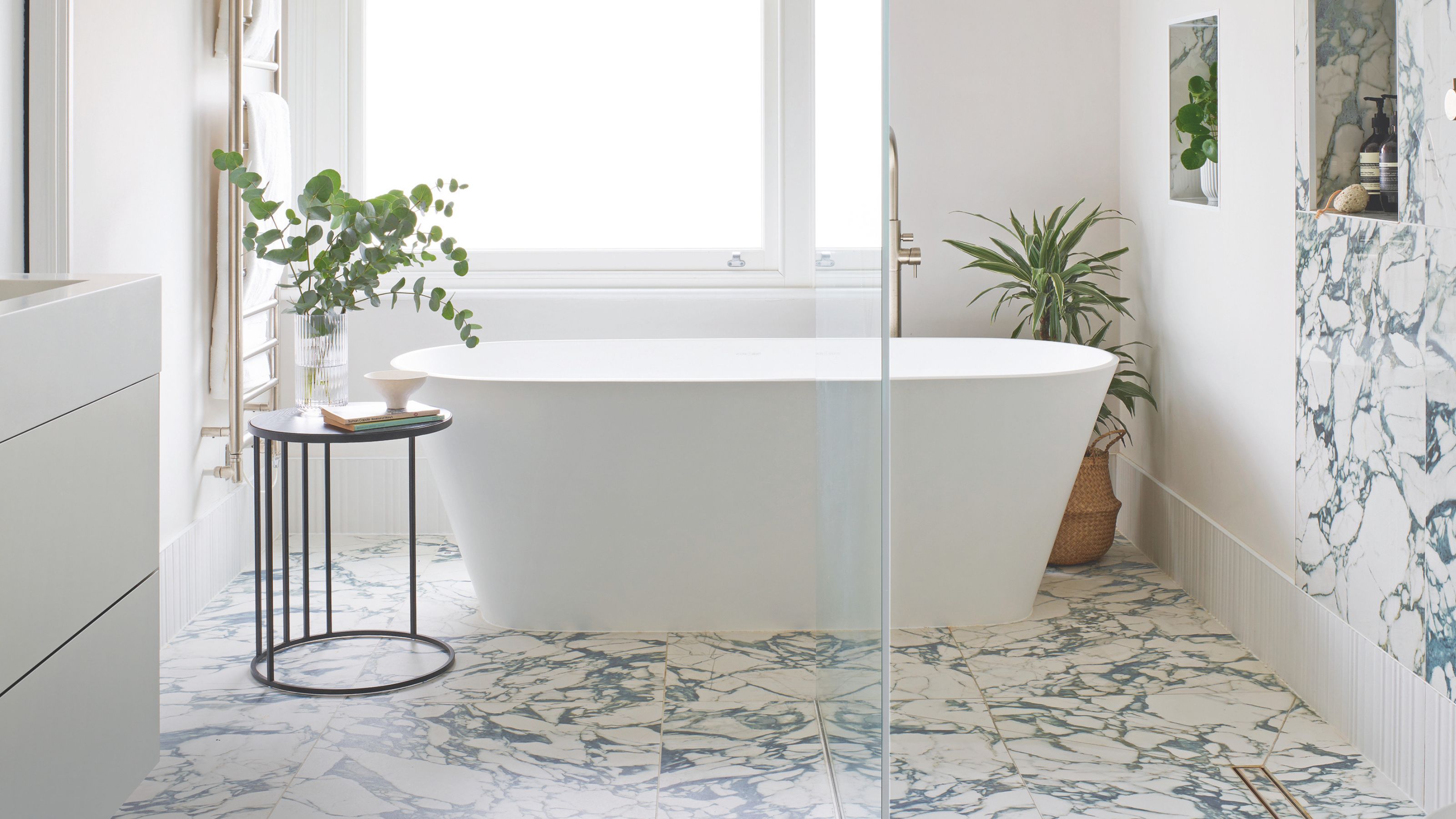 Elegant neutral bathroom with freestanding bath, plants and marble effect floor and wall tiles