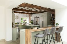 kitchen with exposed wooden beams and grey-green painted cabinets with natural wood breakfast bar and pale stone flooring