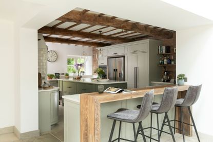 kitchen with exposed wooden beams and grey-green painted cabinets with natural wood breakfast bar and pale stone flooring