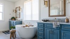 A coastal bathroom with a brown woven large mirror, a gold wall sconce, a blue sink unit with cabinets and drawers and a gray marble surface, gold taps, two oval wooden storage holders and a gray sea shell trinket holder, and a large white freestanding bath with a window above it