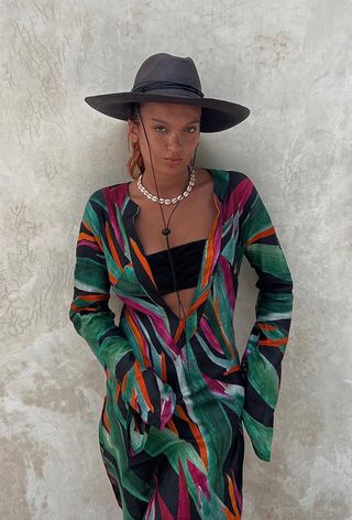 Shell jewelry trend shown on a woman wearing a black fedora with a cowrie shell choker styled with a black bandeau swim top and tropical-print cover-up dress.