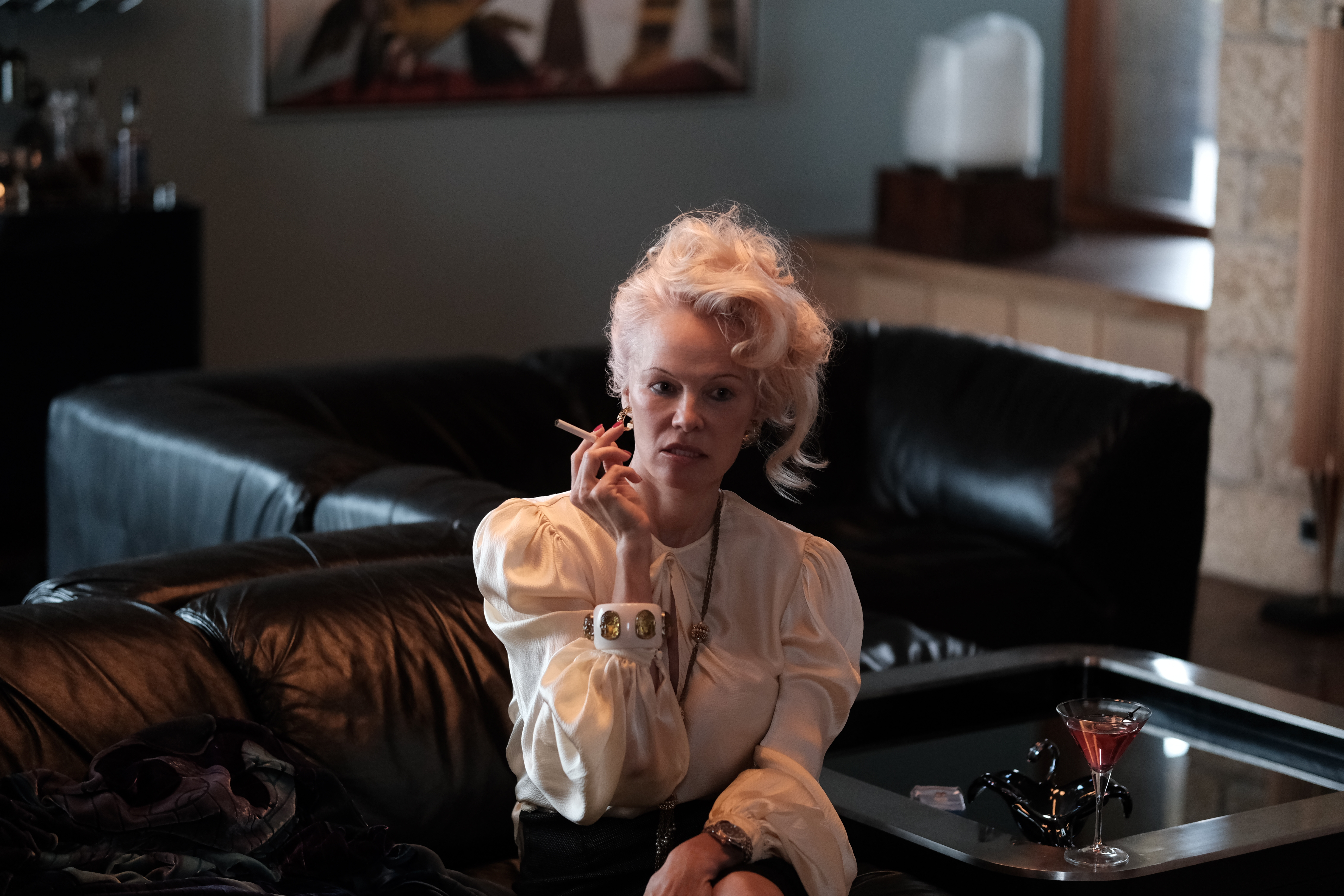 A blonde woman in a voluminous pony tail smokes a cigarette sitting on a black leather sofa while staring to her left.