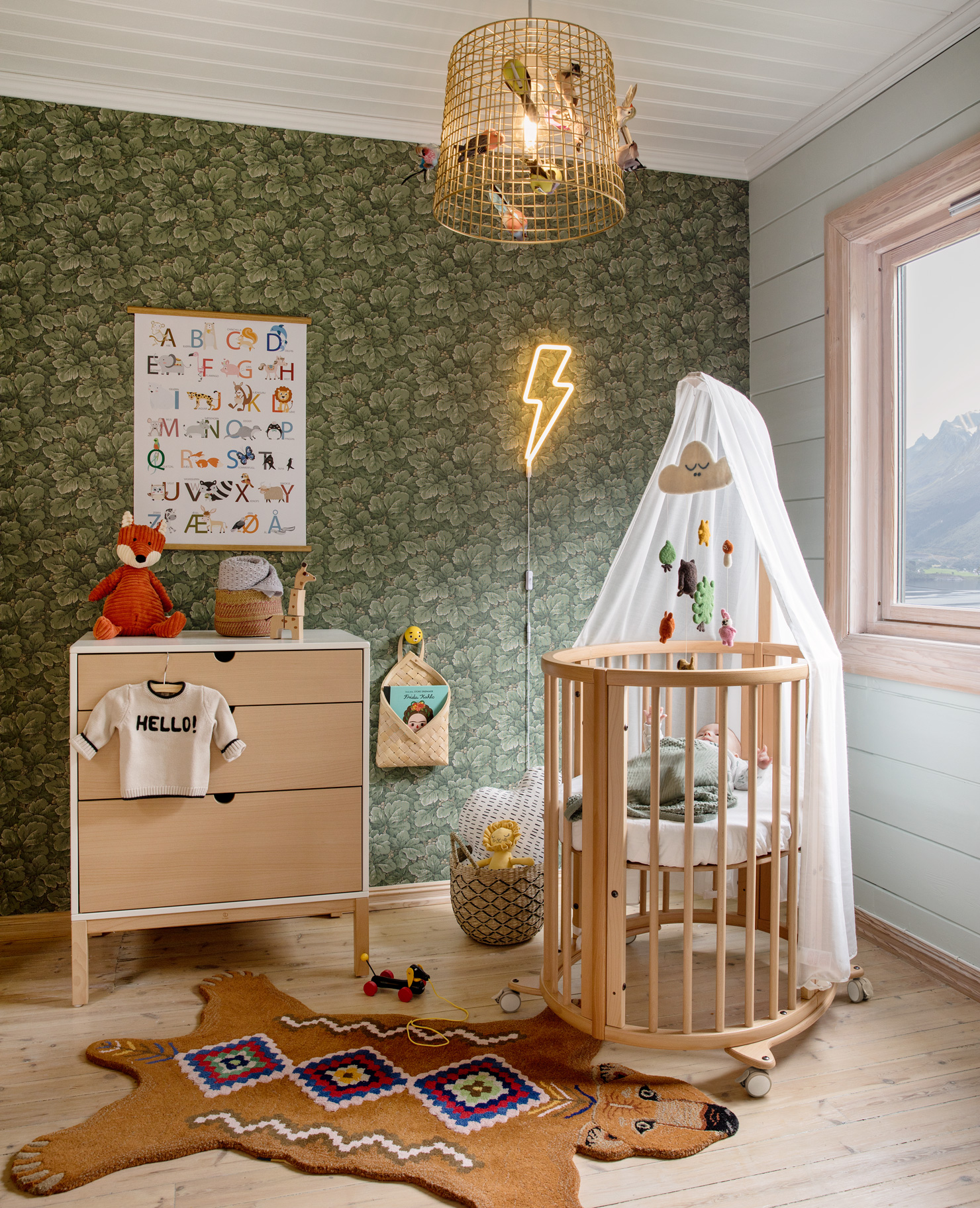 Cot in a coz nursery room with green wallpaper, statement lighting and a faux animal rug.