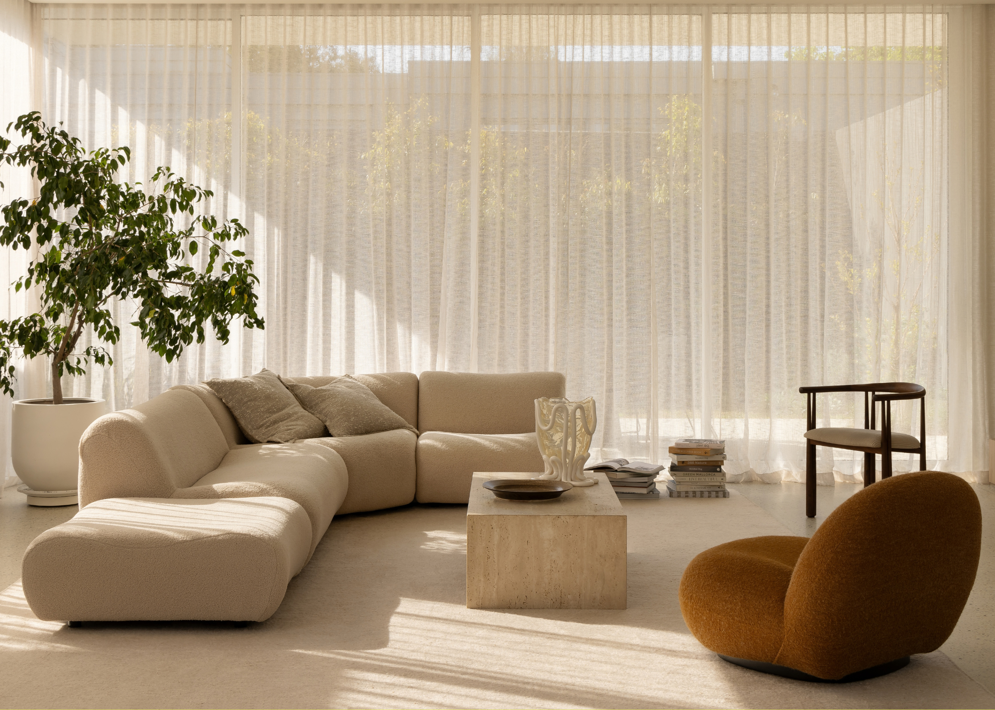 A living room with sheer curtains, a cream sectional sofa, a potted tree, an amber sofa chair, a travertine coffee table and a chair