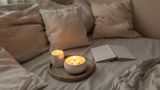 Two lit scented candles on a wooden tray on a bed beside an open book