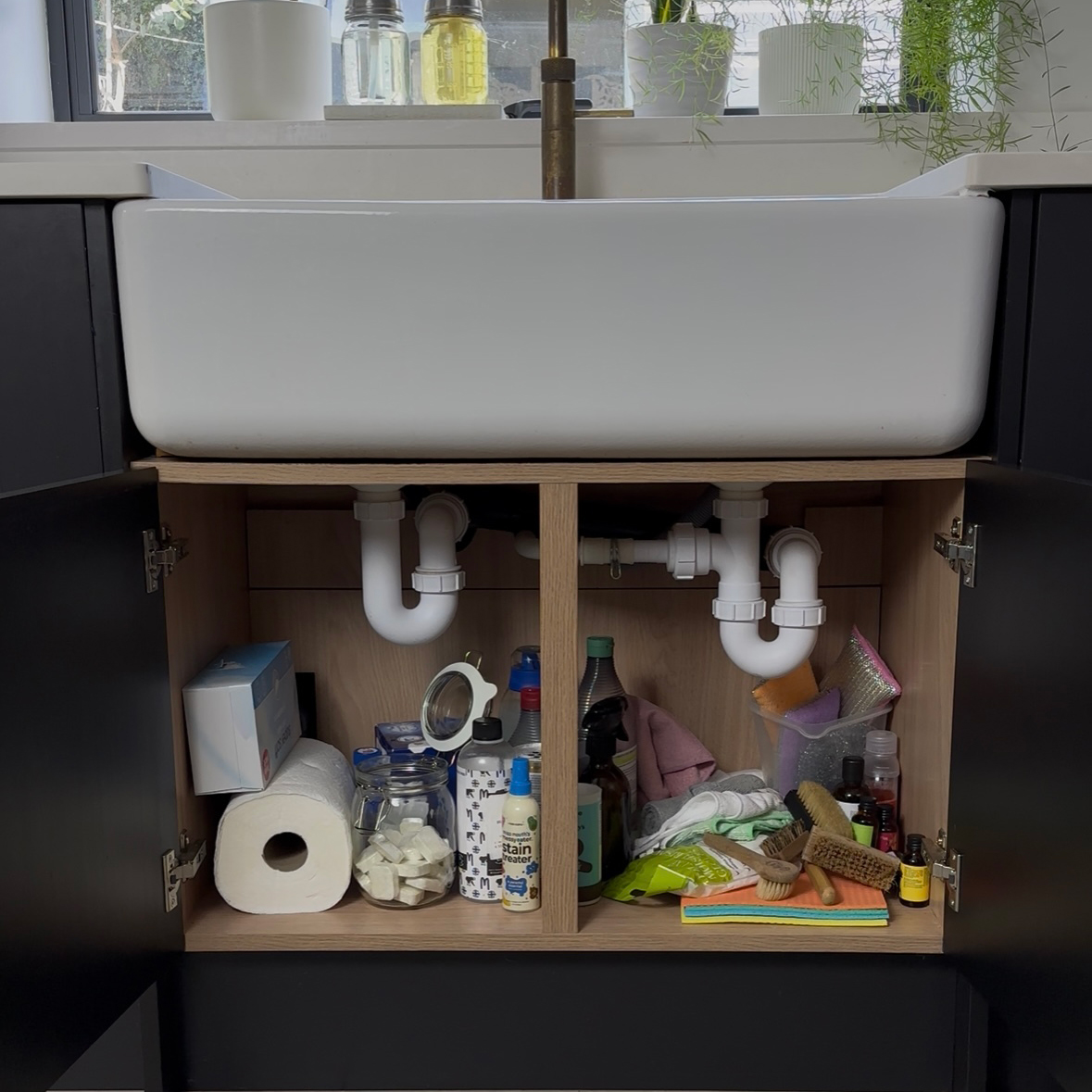 Undersink storage cupboard with doors open showing the interior full of stuff piled up in a disorganised way