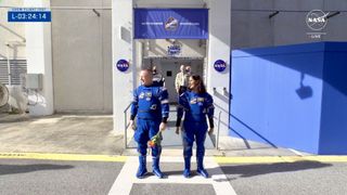 two astronauts in blue spacesuits stand before two pillars with central NASA logos.