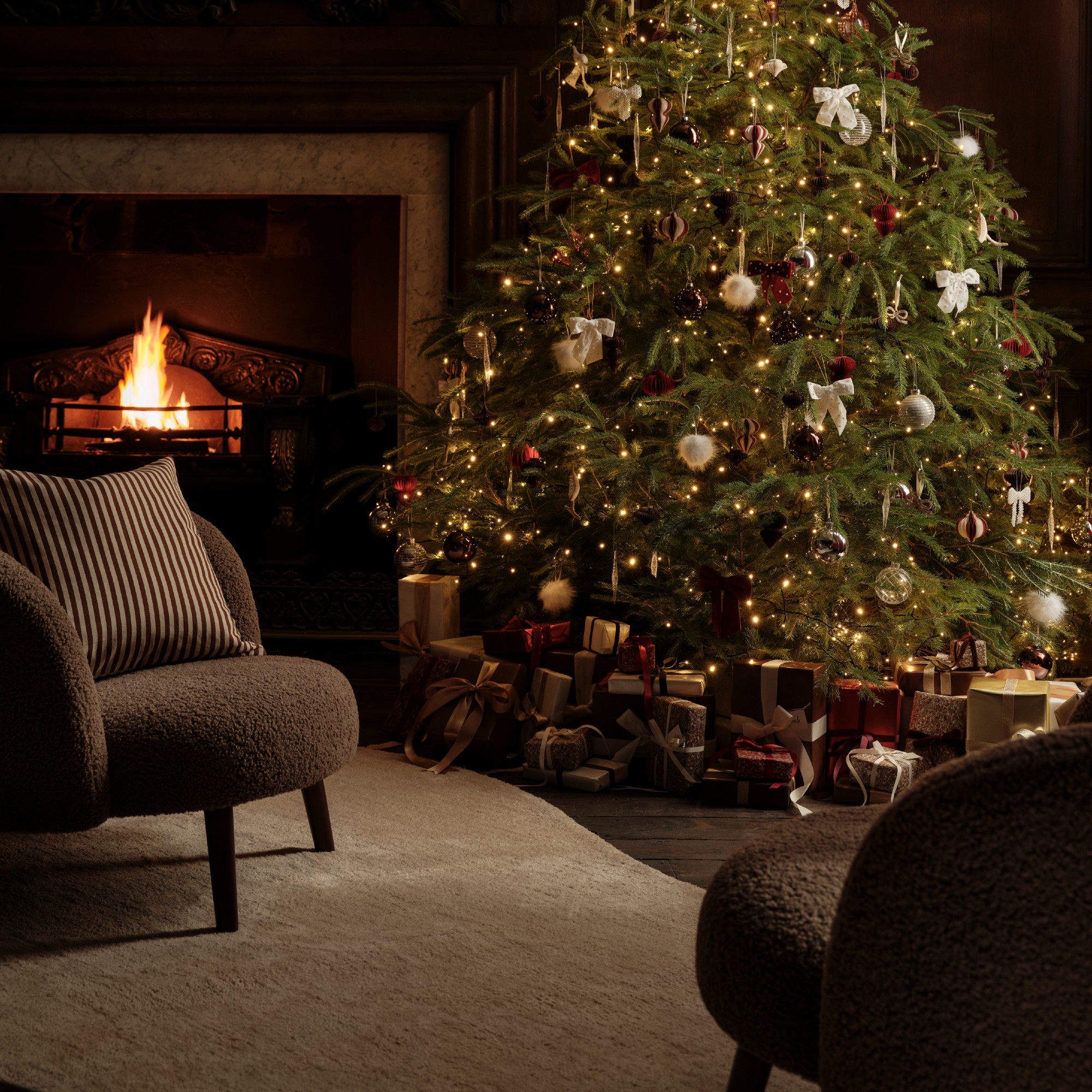 A living room with a decorated lit-up Christmas tree and a lit fireplace with two brown boucle chairs and a striped cushion
