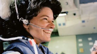 sally ride on flight deck smiling with a headset microphone piece on her head, wearing a blue nasa jumpsuit.