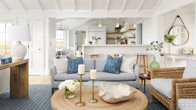 beach house living room with sea views pale blue sofa and accessories round table and mirror with open plan kitchen and dining room in background white walls and ceiling
